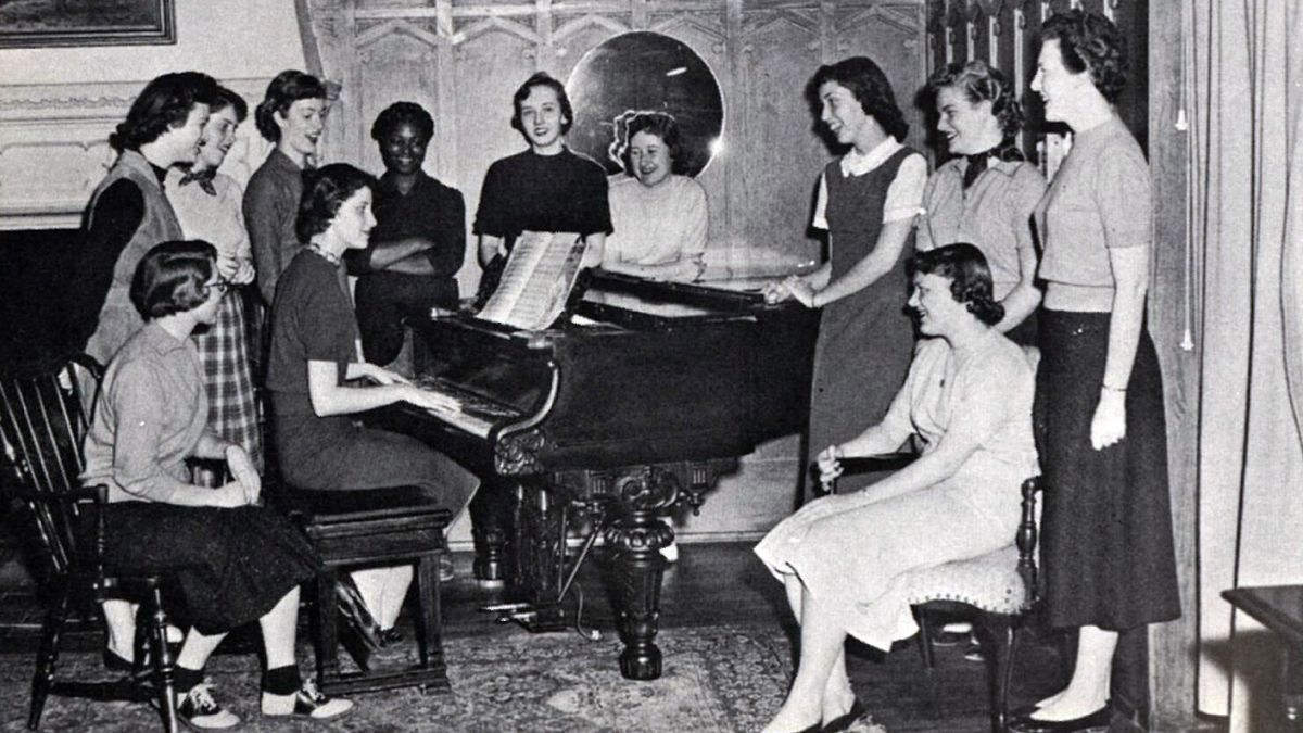 A group of women stand around a piano in South Hall.