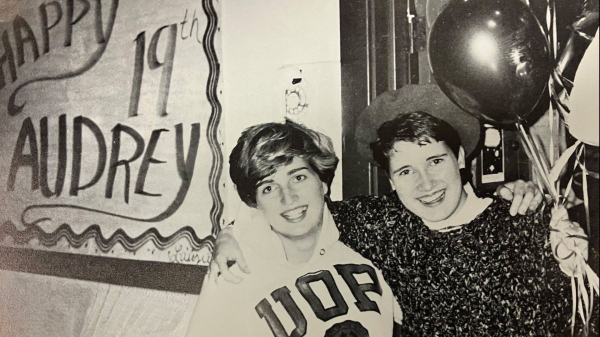 Two friends stand in front of a "Happy Birthday" sign with balloons.