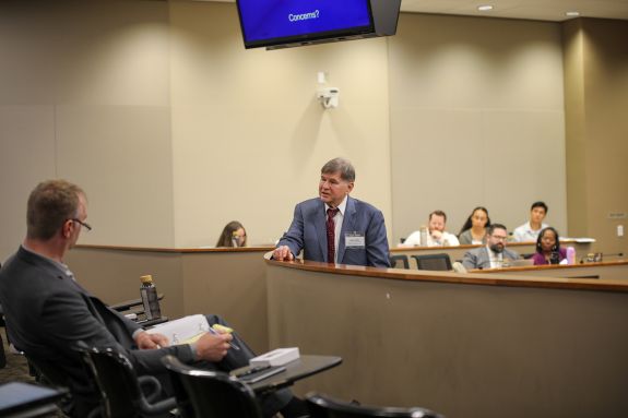 A man stands in front of a crowd of listeners in a courtroom setting. Onlookers can be seen in the background listening attentively. 