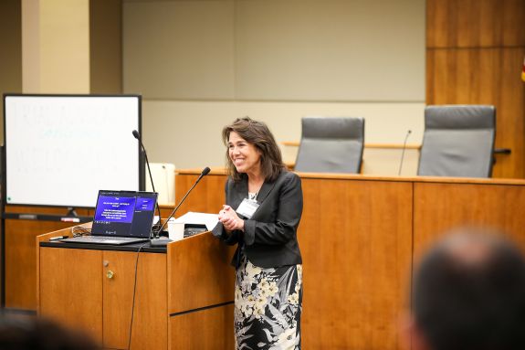 A woman with long dark hair stands in a courtroom with a whiteboard behind her, teaching. The crowd can be seen in the foreground listening. 