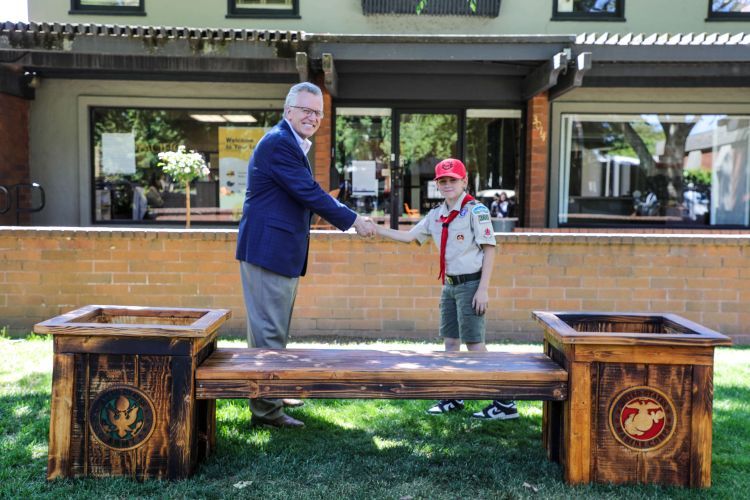 An adult in a blue blazer and khaki pants shakes hands with a child wearing a red cap and scout uniform. They are standing behind a wooden bench with two planters on either side, each bearing an emblem. The background shows a building with large windows and greenery.