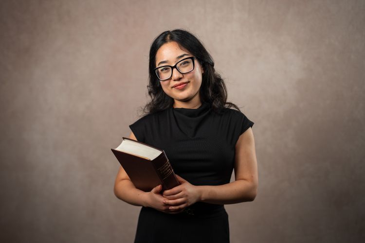 A student holding a textbook in front of a neutral background 