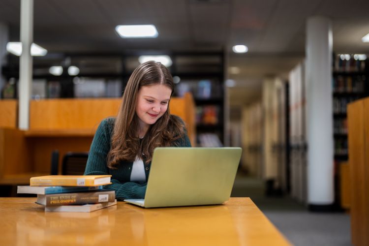 a student sits at a table looking at a laptop