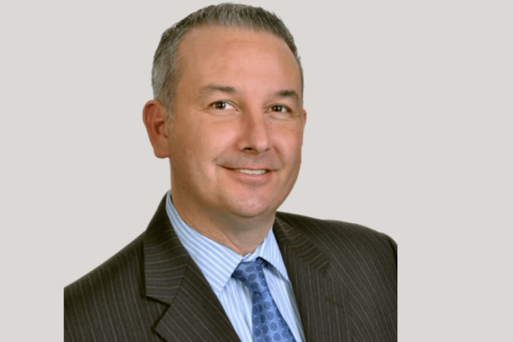 A headshot portrait of a man wearing a grey suit jacket a blue tie and a light blue under shirt. He has short gray hair and brown eyes.  