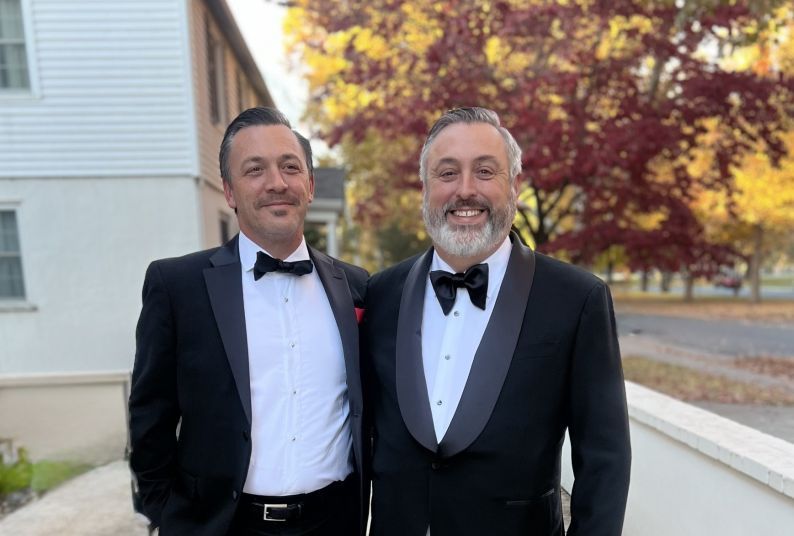 Two brothers stand in an outdoor setting wearing nice suits smiling together. 