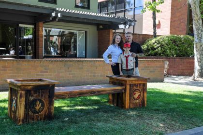 A family of three stands behind a wooden bench with two planters on either side. The bench is located on a grassy area in front of a brick building. The planters have military emblems carved into them.
