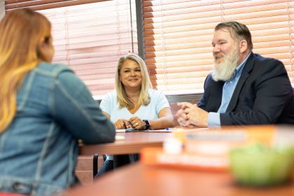 Three people are sitting around a table in a room with wooden blinds. One person is wearing a denim jacket and has long hair, another is in a light blue shirt, and the third is in a suit with a blue shirt. They appear to be engaged in conversation