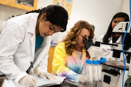 A student in a white lab coat writes in a notebook.