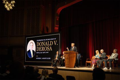 several people sit on a stage next to a large screen that reads "Celebrating the Life and Legacy of Donald V. DeRosa".