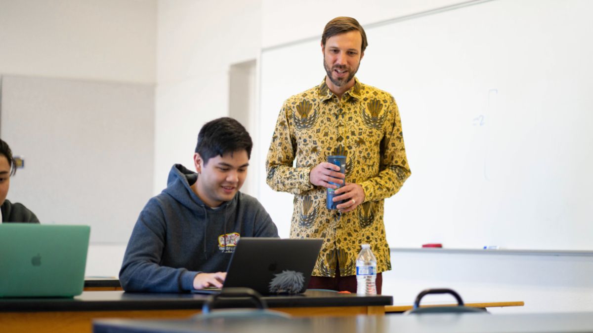 A professor stands next to tables where students work on laptops